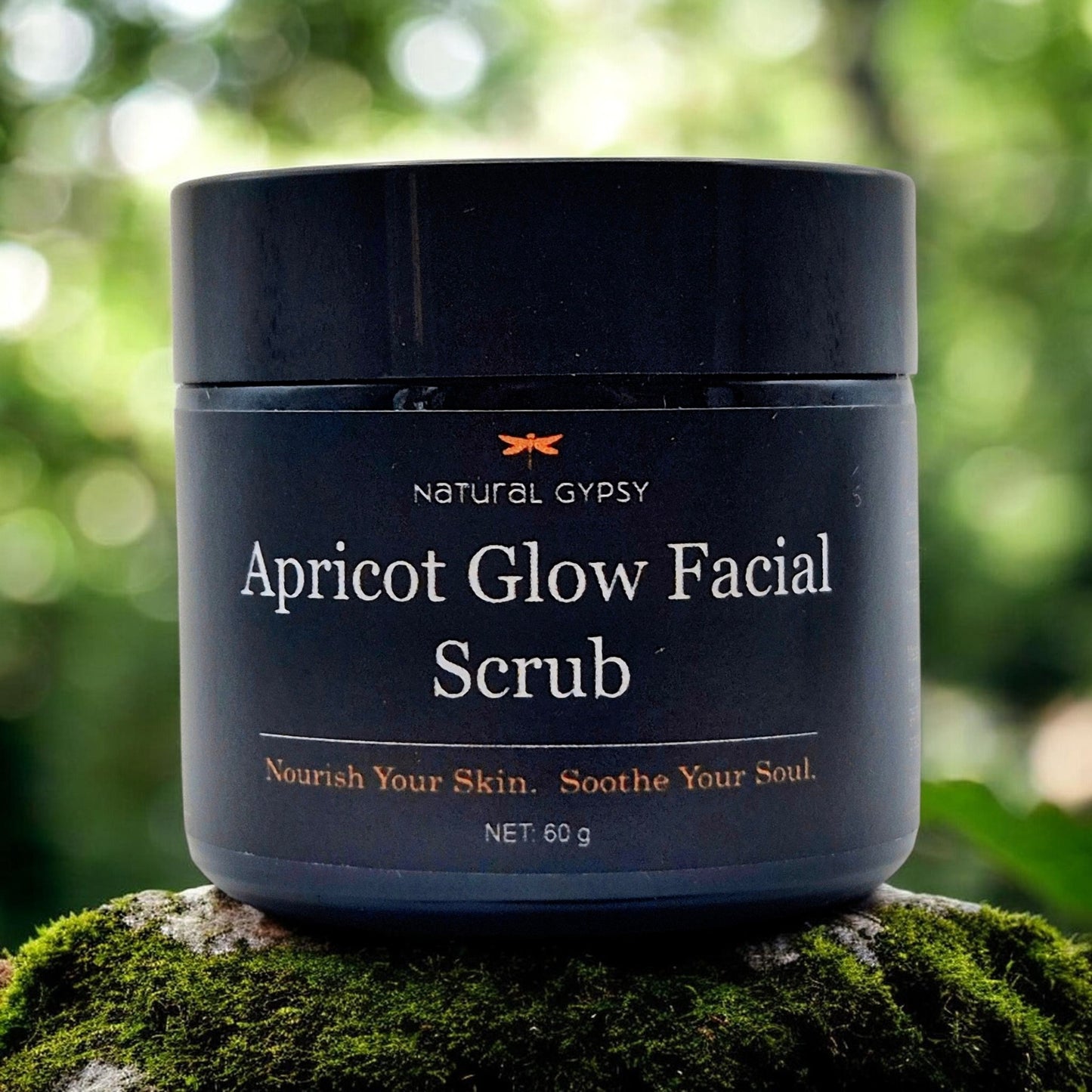 Natural Gypsy Apricot Glow Facial Scrub in a 60g black jar, displayed on a mossy rock in a natural outdoor setting.