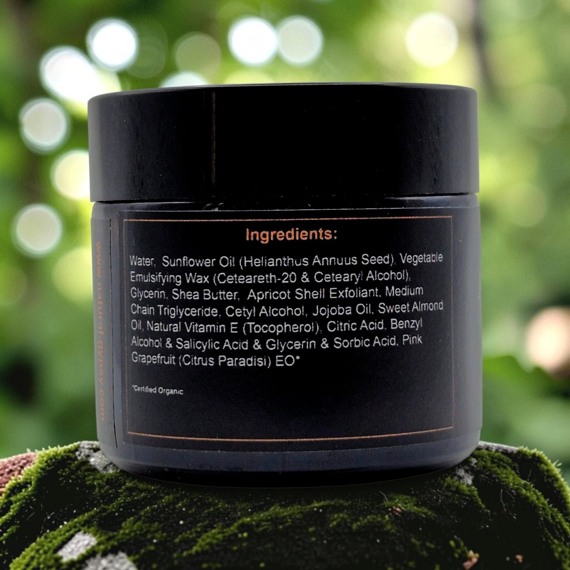 Natural Gypsy Apricot Glow Facial Scrub black jar with ingredient list label on a mossy rock in a garden setting